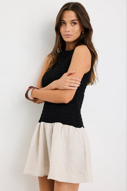 Woman wearing a sleeveless mini dress with a black shirred bodice and a natural beige flared skirt, styled simply for a casual chic look.