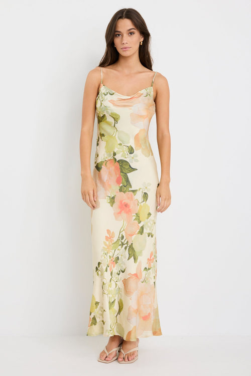 Woman wearing a cream bias-cut slip dress with peach floral print, featuring thin straps, a cowl neckline, and tie-back detail, styled with minimal accessories for a romantic look.