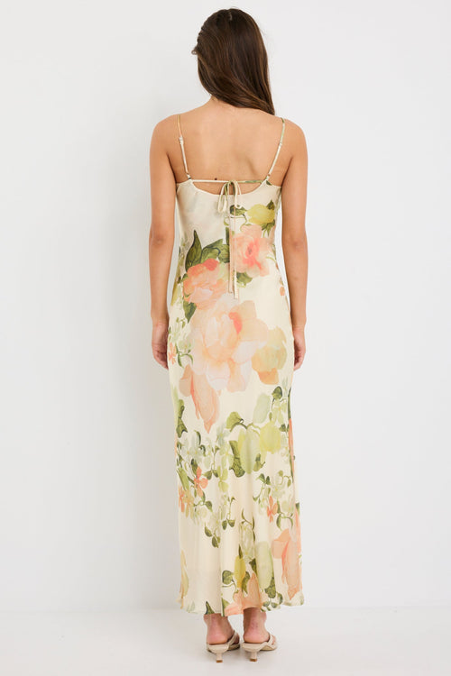 Woman wearing a cream bias-cut slip dress with peach floral print, featuring thin straps, a cowl neckline, and tie-back detail, styled with minimal accessories for a romantic look.