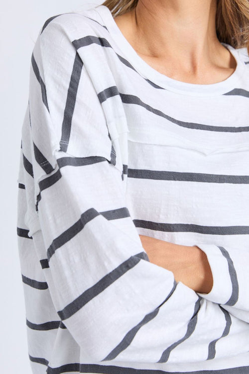 Woman wears a white long sleeve top with black horizontal stripes, crew neck, exposed stitching, and a relaxed fit. Styled with blue jeans for a casual, easy-going look.