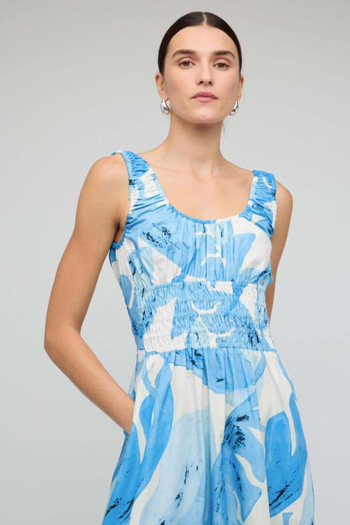 Woman wearing a light blue and white sleeveless midi dress with a shirred waist and soft leaf print, featuring a relaxed, floaty silhouette.