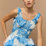 Woman wearing a light blue and white sleeveless midi dress with a shirred waist and soft leaf print, featuring a relaxed, floaty silhouette.