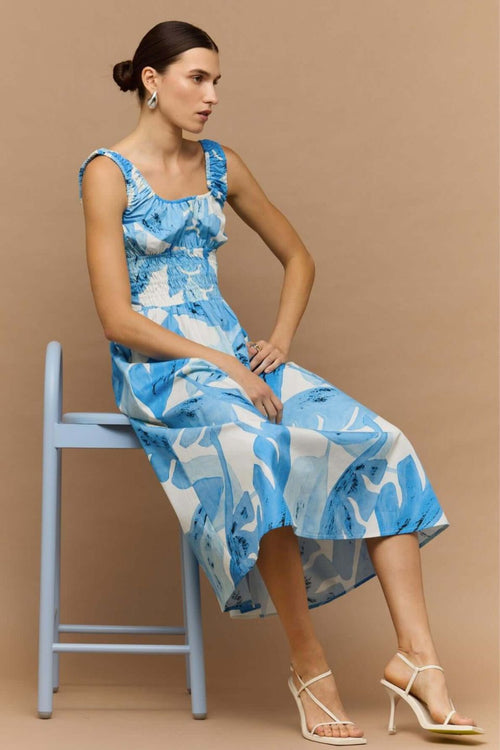 Woman wearing a light blue and white sleeveless midi dress with a shirred waist and soft leaf print, featuring a relaxed, floaty silhouette.