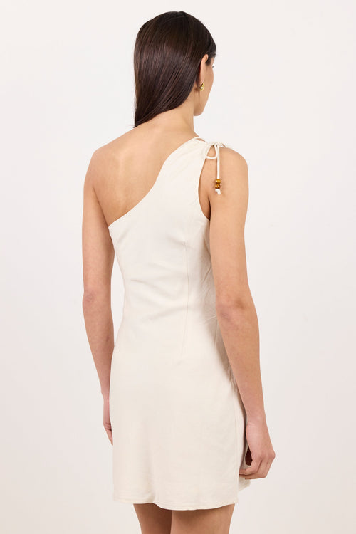 A woman viewed from the back wearing a one-shoulder cream dress with a small gold tassel detail on the strap, set against a plain off-white studio background.