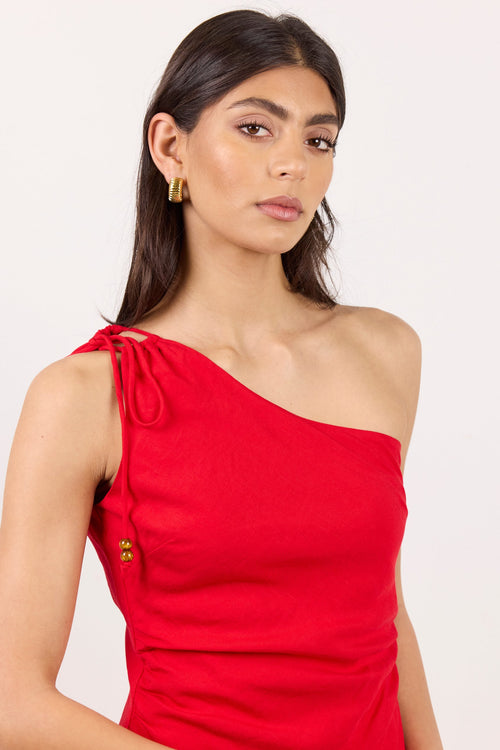 A woman modeling a bright red one-shoulder wrap dress with a tie detail on the shoulder and a small gold button accent, set against a plain light background.