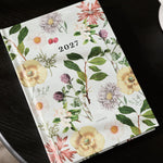 A 2027 floral planner or desk calendar lies open on a dark round table, showing a botanical cover with yellow flowers, green leaves, and small pink and purple blossoms. A green ribbed candle holder and part of a white object are visible in the background.