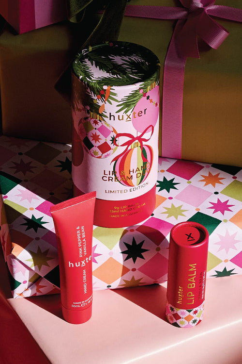 A huxter Lip balm set in a gift-wrapped environment, featuring a cylindrical tin of Lip Happy Cream, a red tube of Lip Balm, and a red stick of Lip Balm on a pink surface with colorful gift boxes in the background.