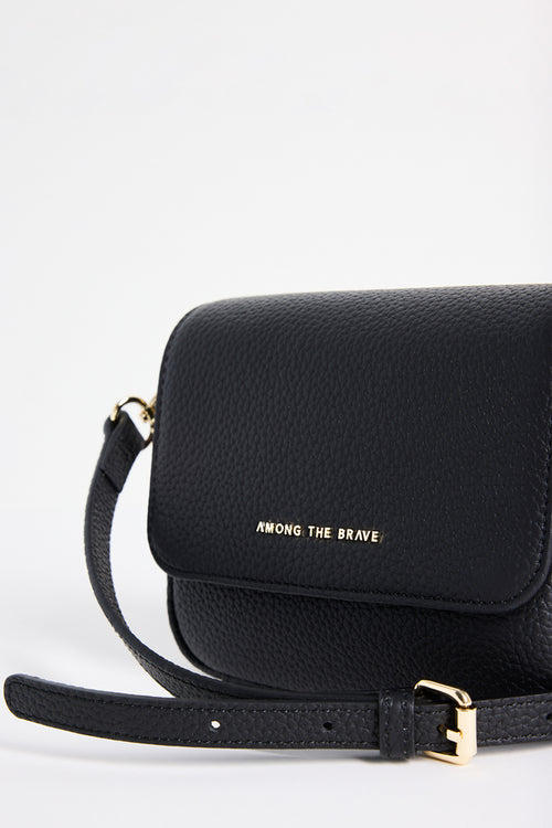 A close-up of a black pebbled leather crossbody bag with gold lettering that reads 'AMONG THE BRAVE' on the front flap. The bag features a black leather strap with gold-tone hardware, set against a clean white background.