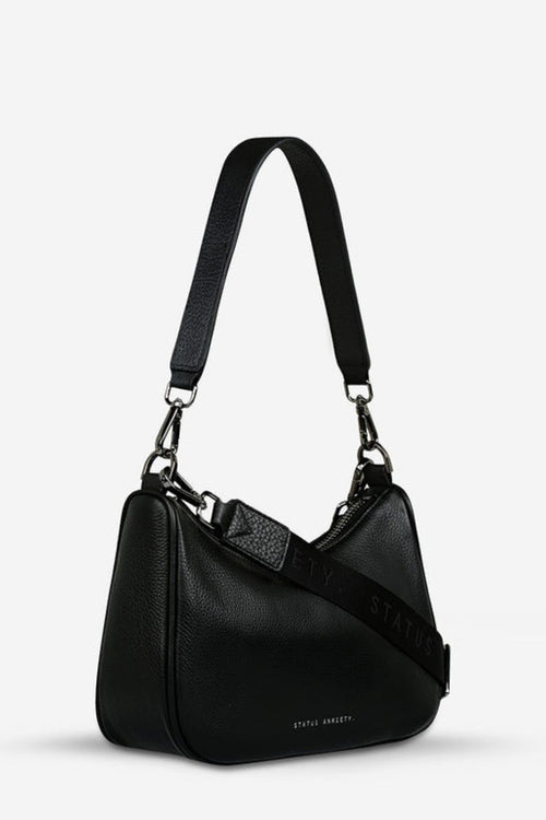 A sleek black leather crossbody bag with a soft, curved silhouette and pebble-grain texture. It features a detachable shoulder strap with silver hardware clasps and 