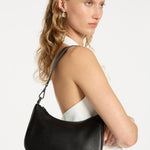 A sleek black leather crossbody bag with a soft, curved silhouette and pebble-grain texture. It features a detachable shoulder strap with silver hardware clasps and "Status Anxiety." branding subtly printed in white on the lower front.