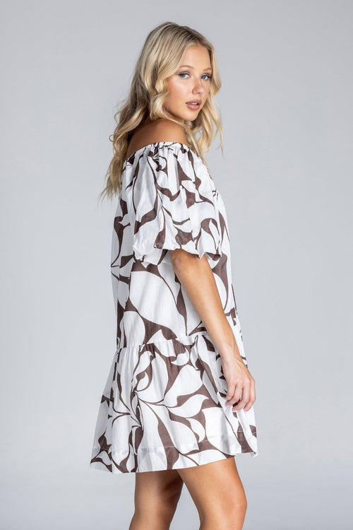 Woman wearing an off-the-shoulder white mini dress with abstract brown swirl print, puff sleeves, and a tiered skirt, standing against a plain grey background.