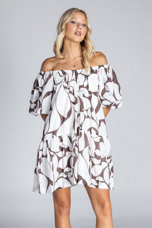 Woman wearing an off-the-shoulder white mini dress with abstract brown swirl print, puff sleeves, and a tiered skirt, standing against a plain grey background.
