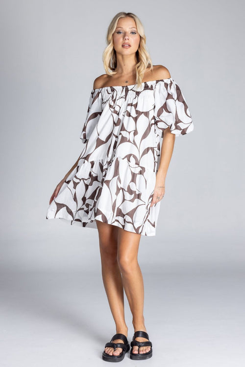 Woman wearing an off-the-shoulder white mini dress with abstract brown swirl print, puff sleeves, and a tiered skirt, standing against a plain grey background.