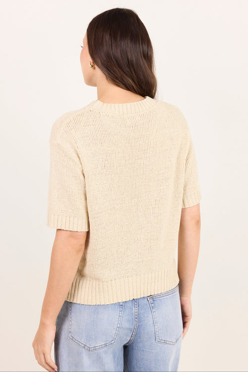 A woman viewed from the back wearing a short-sleeve cream knit sweater and light blue jeans against a plain off-white studio background.