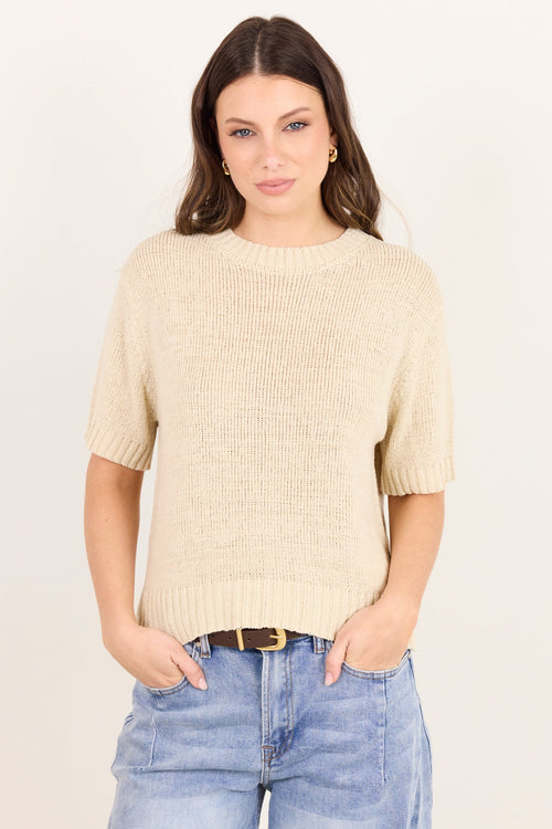 A woman modeling a short-sleeve cream-colored ribbed knit sweater with light-wash blue jeans against a plain off-white background.