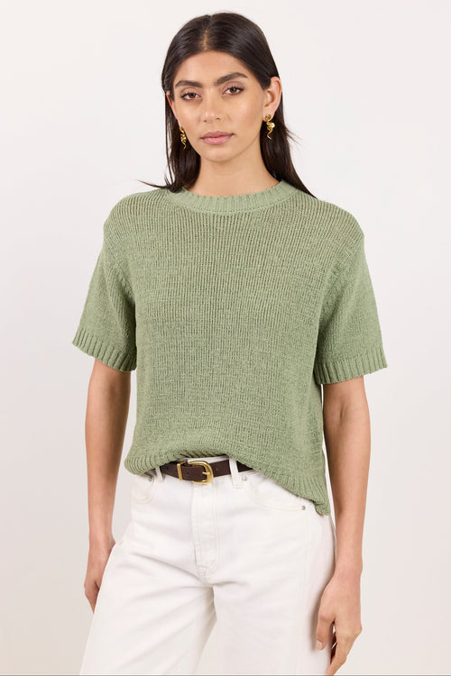 A woman modeling a short-sleeve ribbed knit sweater in sage green, paired with white pants and a brown belt, against a plain off-white studio background.
