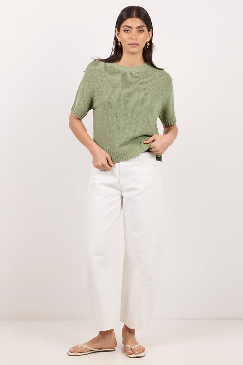 A woman modeling a short-sleeve sage green knit top with white cropped pants and flat sandals, standing against a plain white studio background.