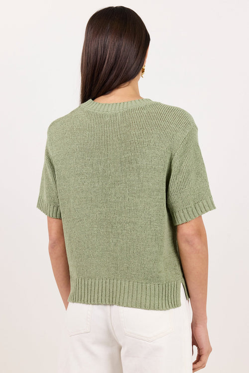 A woman viewed from the back wearing a short-sleeve knit sweater in a muted sage green and white pants, against a plain off-white studio background.