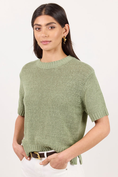 A woman modeling a short-sleeve knit sweater in sage green, paired with white pants and a brown belt, against a plain white studio background.