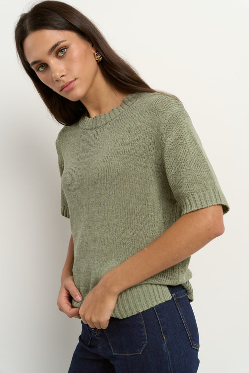 Woman wearing a short sleeve sage knit top with ribbed edges and light blue jeans.