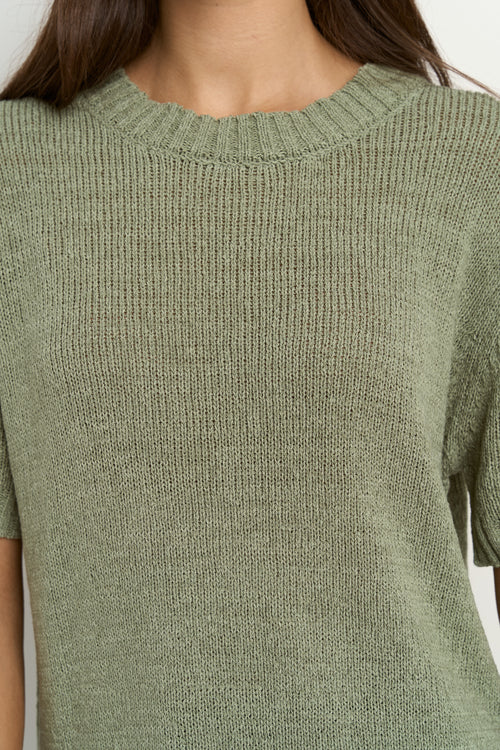 Woman wearing a short sleeve sage knit top with ribbed edges and light blue jeans.