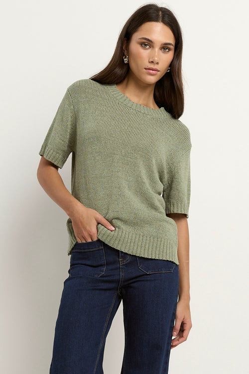 Woman wearing a short sleeve sage knit top with ribbed edges and light blue jeans.
