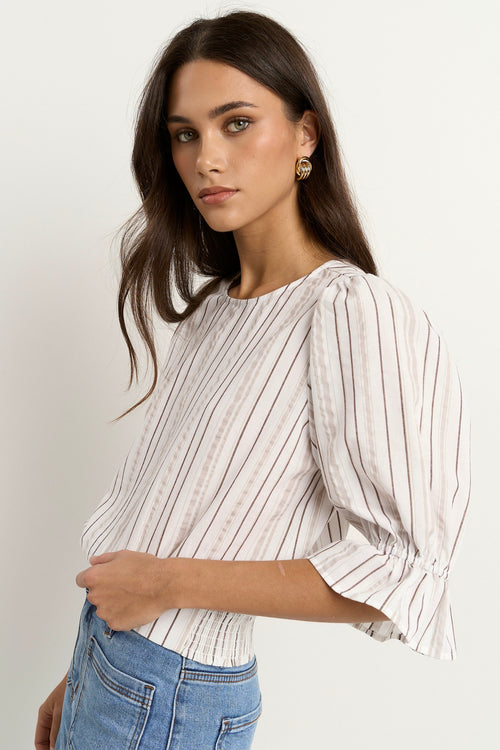 Woman wearing a puff-sleeve chocolate stripe top with shirred waist and light denim jeans.