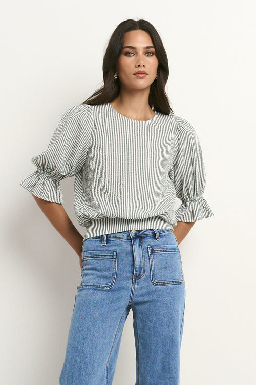 Woman wears a khaki and white striped puff sleeve top with frill detail, paired with light denim jeans.