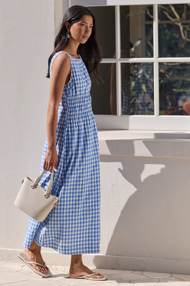 A woman wearing a blue and white gingham sleeveless maxi dress walks outdoors next to a white wall with a window, holding a light beige handbag.