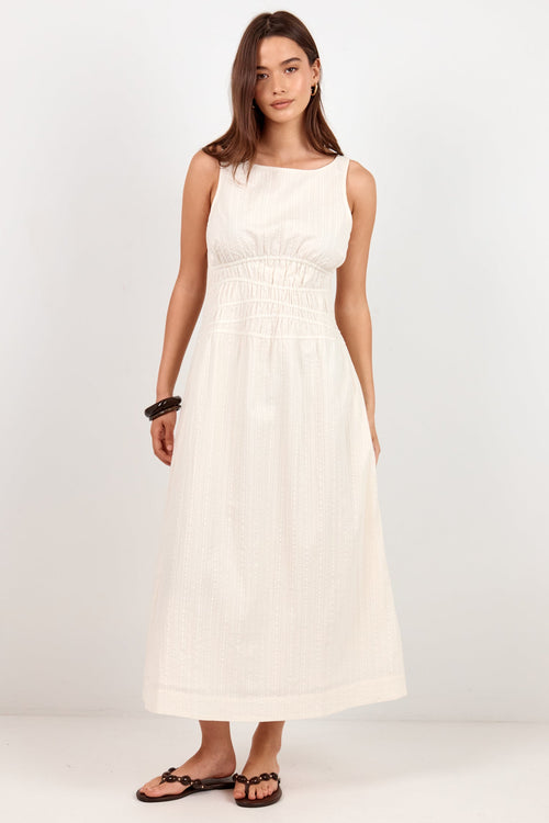 A woman modeling a sleeveless white midi dress with a smocked bodice and gathered skirt, paired with black flat sandals, against a plain white studio background.