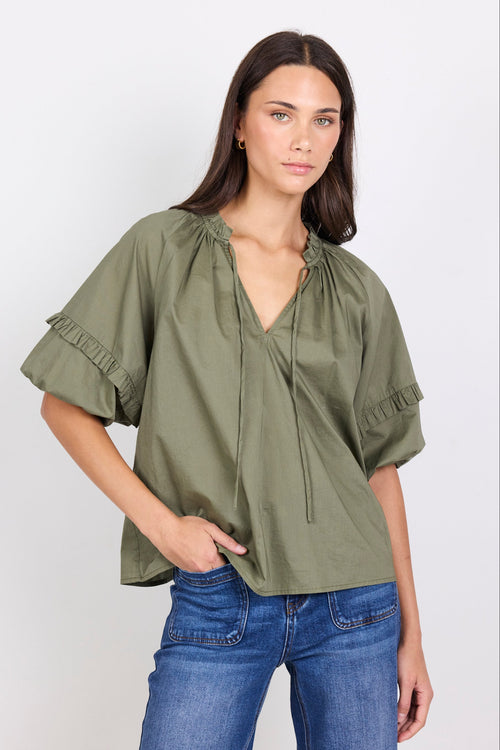 Woman wearing an olive green blouse with ruffled sleeves and blue jeans on a white background