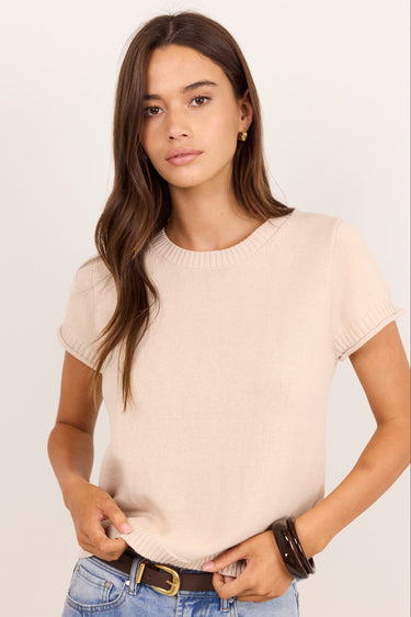 A woman modeling a short-sleeve crewneck knit top in a light beige color, paired with blue jeans and a brown belt, against a plain light background.