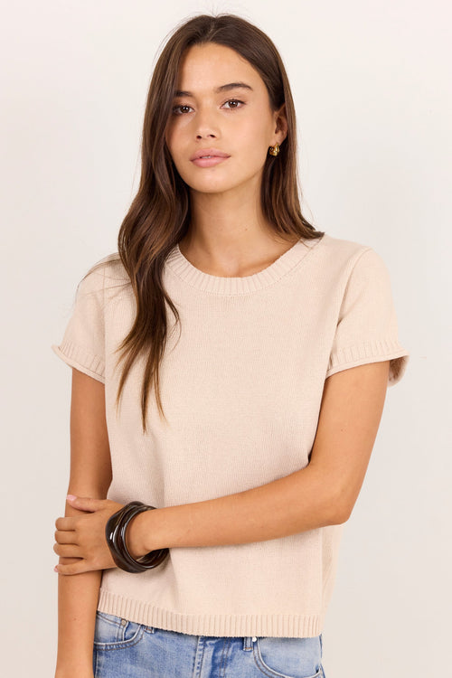 A woman modeling a short-sleeve crewneck knit top in a light beige color, paired with blue jeans, against a plain off-white studio background.