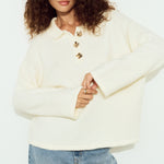 Woman wearing a cream sweater with gold buttons and blue jeans on a white background