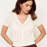 A woman wearing a cream short-sleeve knit top with a V-neck and brown wide-leg pants with white polka dots, posing against a plain off-white background.