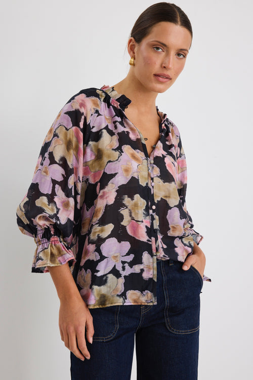 Woman wearing a loose-fit floral blouse in a black base with pink, lilac and beige blooms, featuring ruffled collar and cuffs, styled with dark denim jeans.