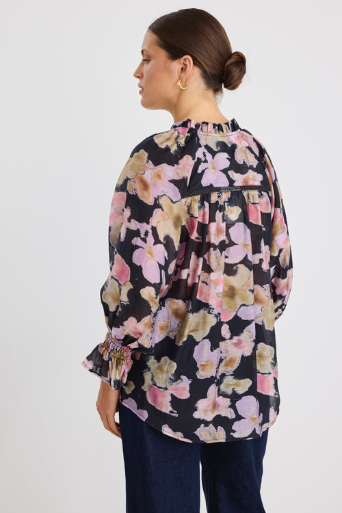 Woman wearing a loose-fit floral blouse in a black base with pink, lilac and beige blooms, featuring ruffled collar and cuffs, styled with dark denim jeans.