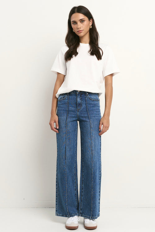Woman wearing high rise, wide leg classic blue jeans with front seam detail, styled with a white tee and white sneakers.