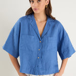 Woman wearing an oversized short-sleeve cornflower blue linen shirt with button front and chest pockets, styled with ecru drawstring jeans.