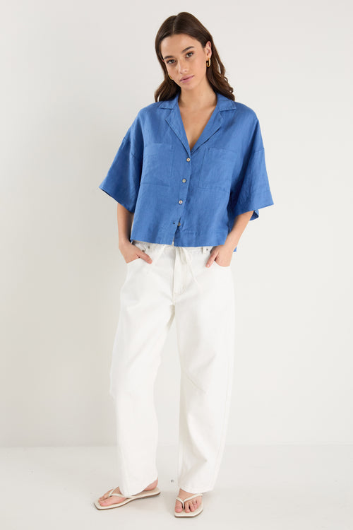 Woman wearing an oversized short-sleeve cornflower blue linen shirt with button front and chest pockets, styled with ecru drawstring jeans.