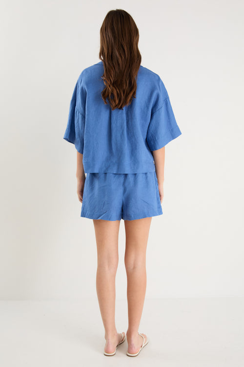 Woman wearing high-rise cornflower blue linen pull-on shorts with rope tie detail, styled with a plain ivory tee.