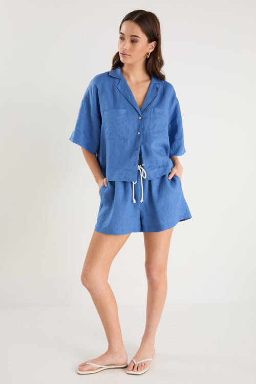 Woman wearing an oversized short-sleeve cornflower blue linen shirt with button front and chest pockets, styled with ecru drawstring jeans.