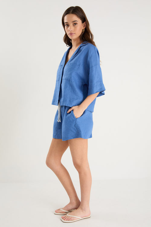 Woman wearing high-rise cornflower blue linen pull-on shorts with rope tie detail, styled with a plain ivory tee.