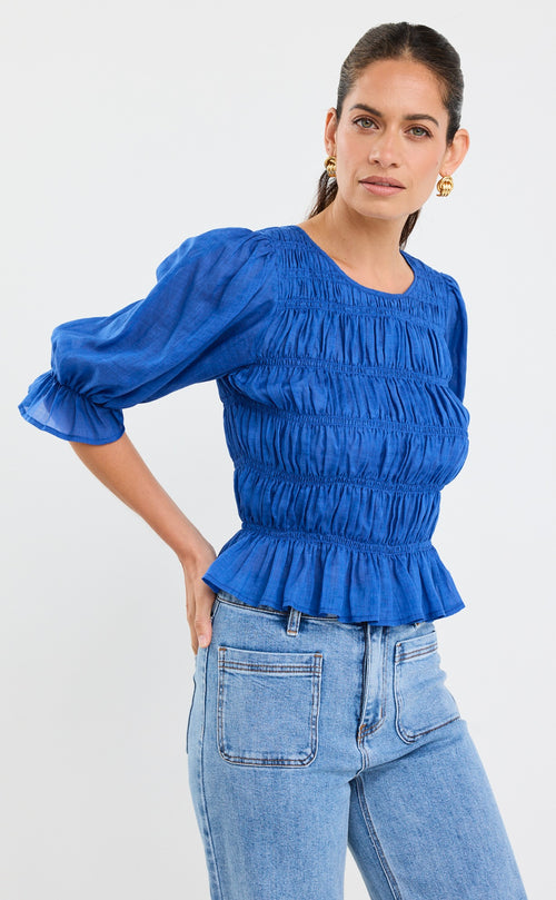 Woman wearing a bright blue ruched cotton top with puff sleeves and peplum hem, styled with light blue jeans.