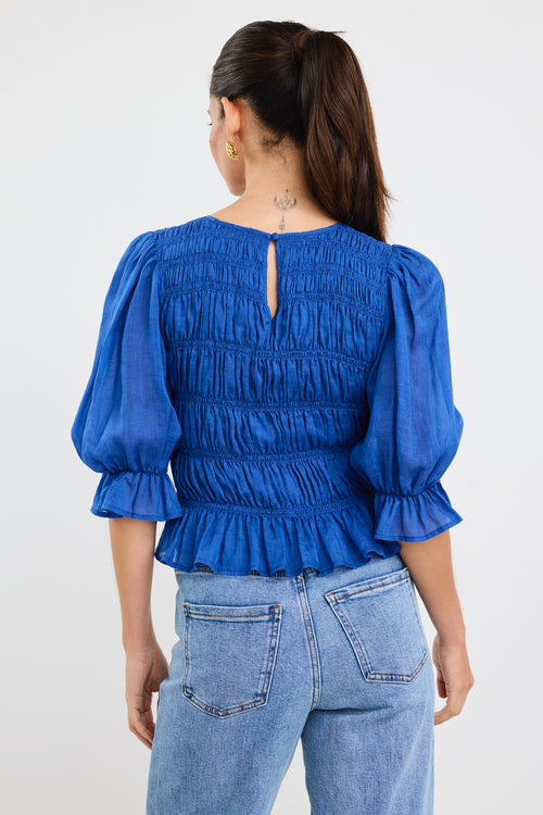 Woman wearing a bright blue ruched cotton top with puff sleeves and peplum hem, styled with light blue jeans.