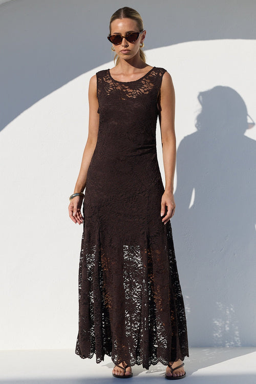 A woman modeling a sleeveless, floor-length dark brown lace dress in front of a white arched wall, casting a long shadow. She is wearing dark sunglasses and holding a small black clutch.