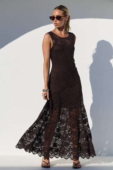 A woman modeling a sleeveless, floor-length dark brown lace dress with a fitted bodice and flared skirt, standing in front of a white arched wall with a strong shadow.