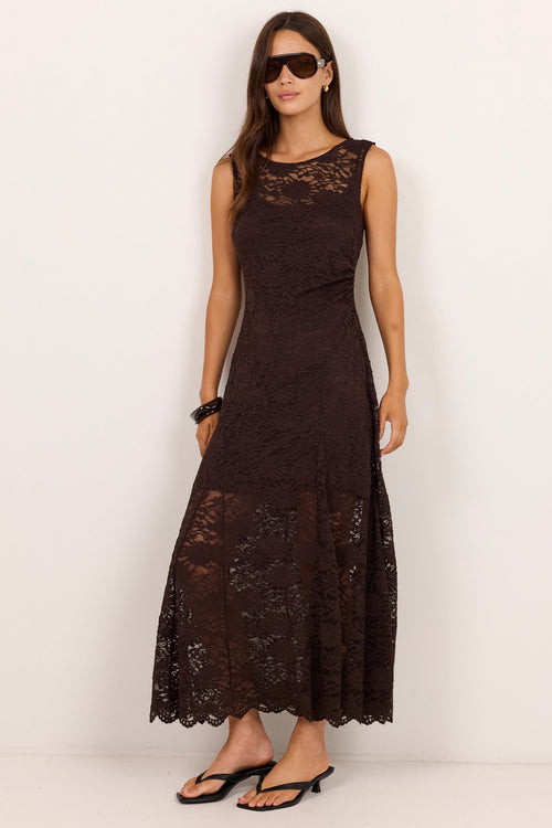 A woman modeling a sleeveless, dark brown lace maxi dress with black flat sandals against a plain white studio background.