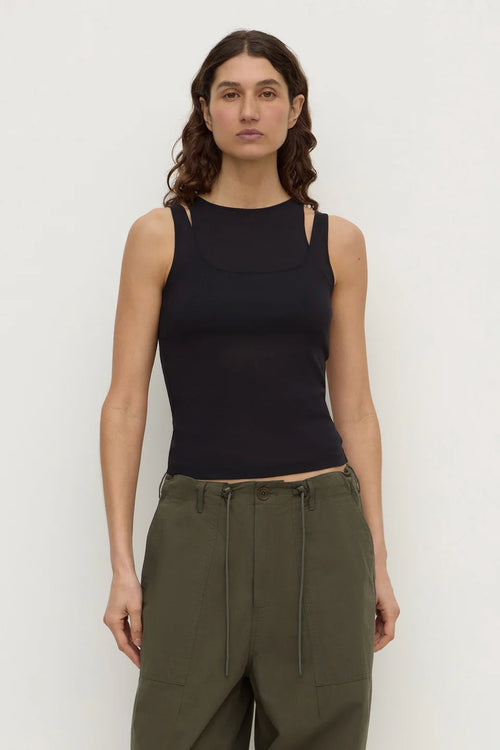 A woman modeling a black sleeveless knit top with cutout details at the shoulders and sides, paired with olive green pants, against a plain light gray studio background.