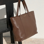 A brown leather tote bag with long shoulder straps, hanging from the backrest of a black wooden chair against a textured off-white wall and light stone floor.